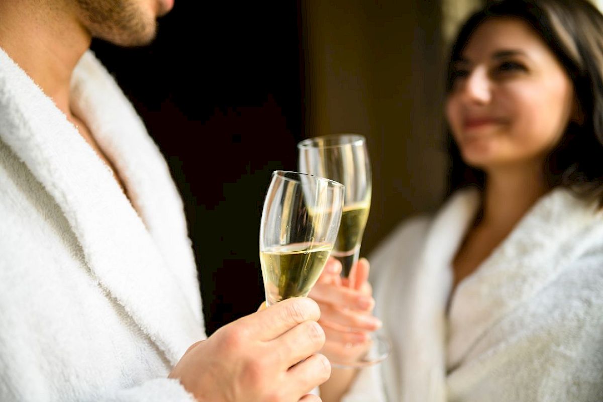 Two people in white robes are toasting with glasses of champagne. The focus is on their hands holding the glasses, with smiles visible.
