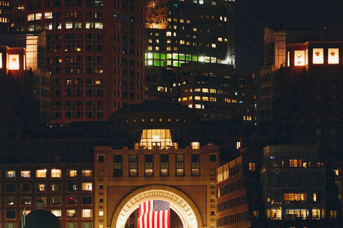 An illuminated cityscape at night features a prominent building with an archway displaying an American flag, reflected on the water's surface.