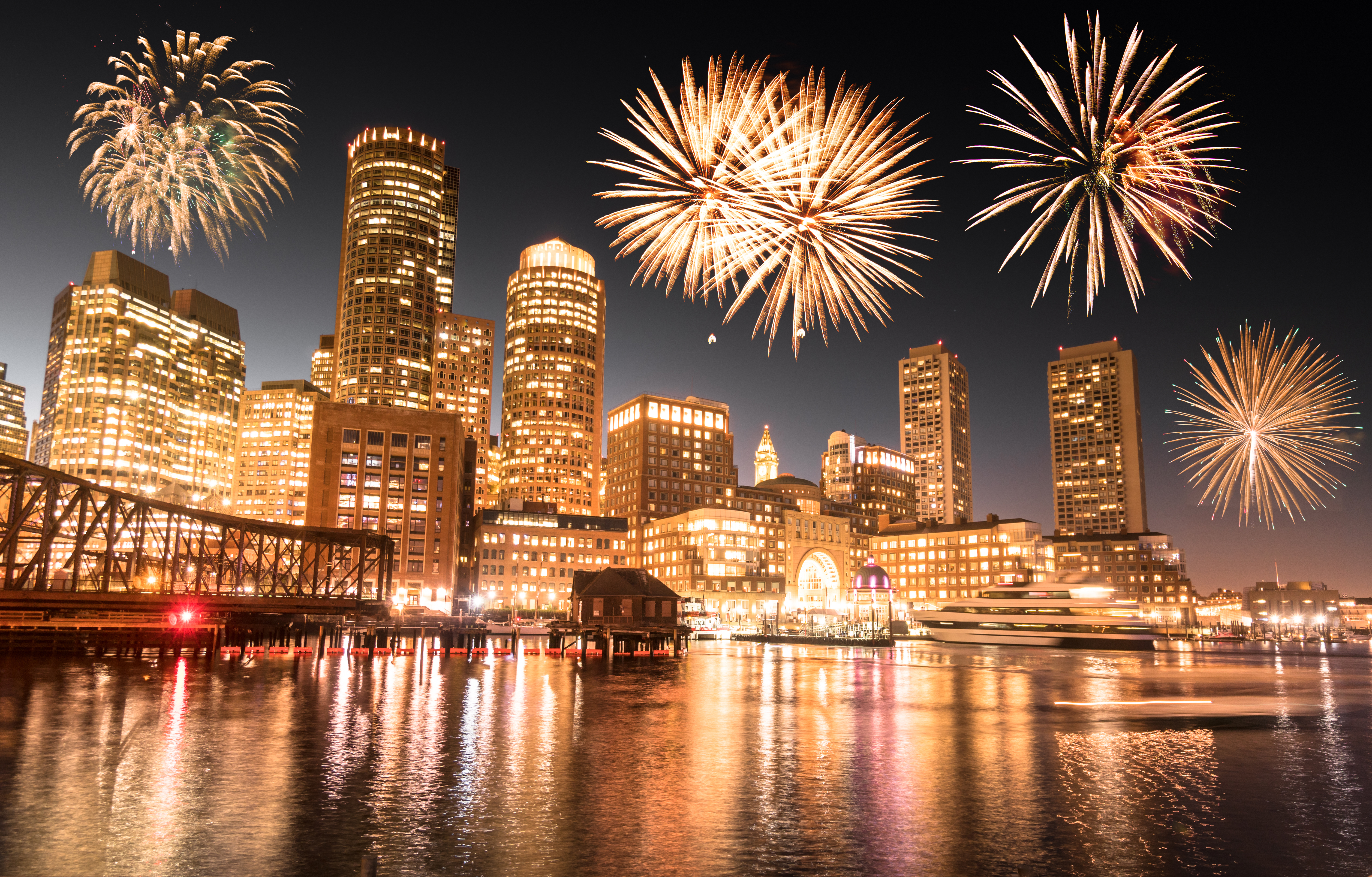 A city skyline at night with fireworks exploding above the harbor and reflected lights on the water.