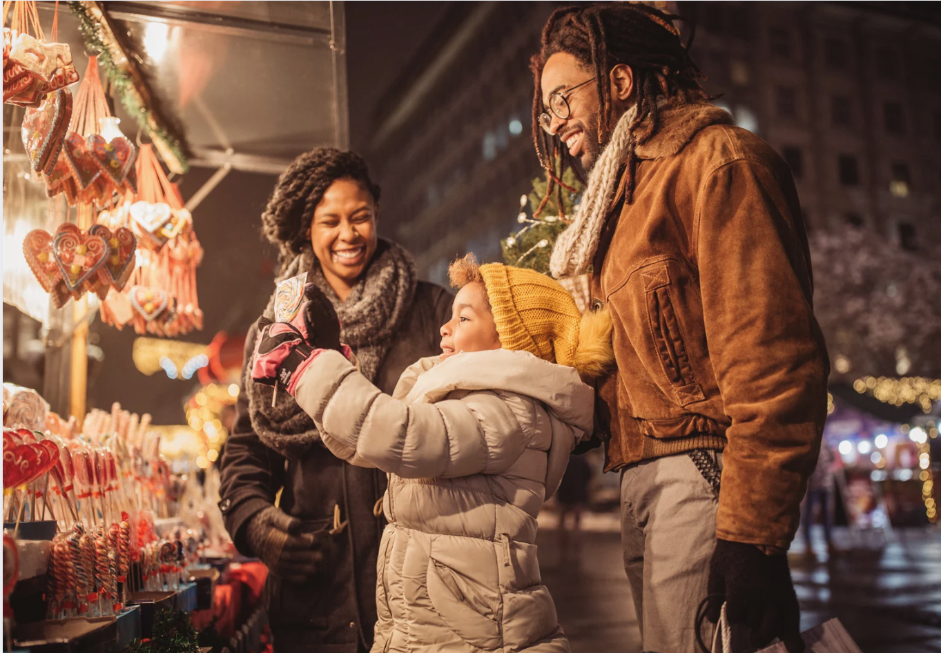 Three smiling adults and a baby at a festive market stall, admiring heart-shaped decorations under twinkling lights, joyful family moment.