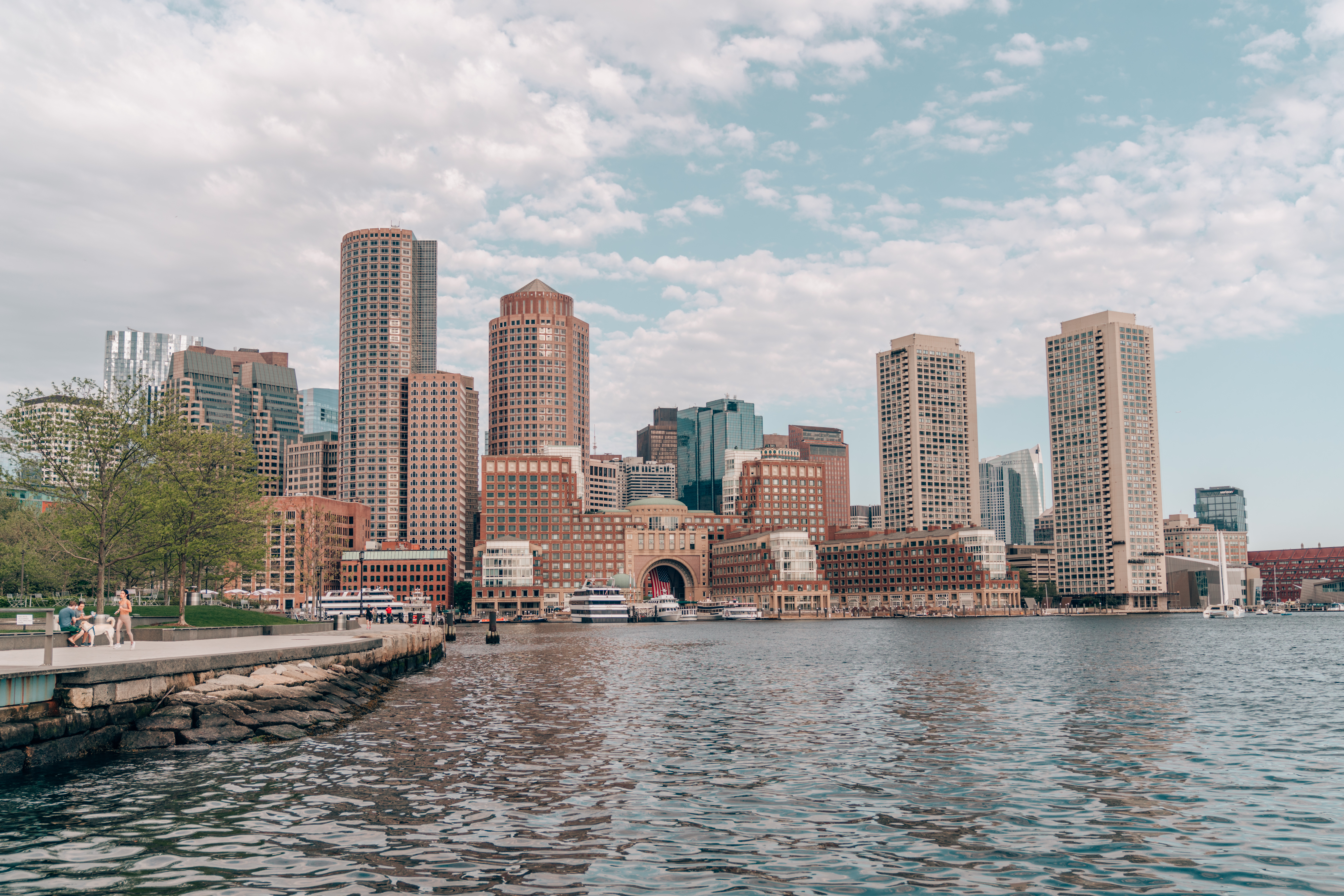 Boston skyline along the harbor with tall buildings, calm water in the foreground, a few boats, and a partly cloudy sky.
