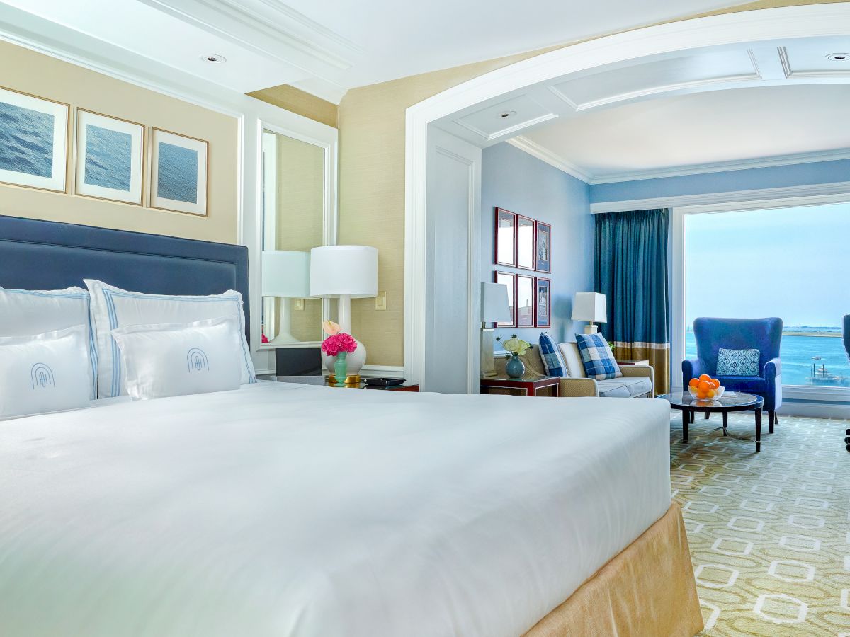 A luxurious hotel room with a large bed, seating area, and ocean view, decorated in light and blue tones with elegant furnishings.