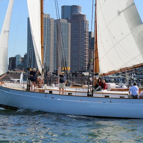 A sailboat with people onboard is sailing in the water, with a cityscape featuring tall buildings in the background.