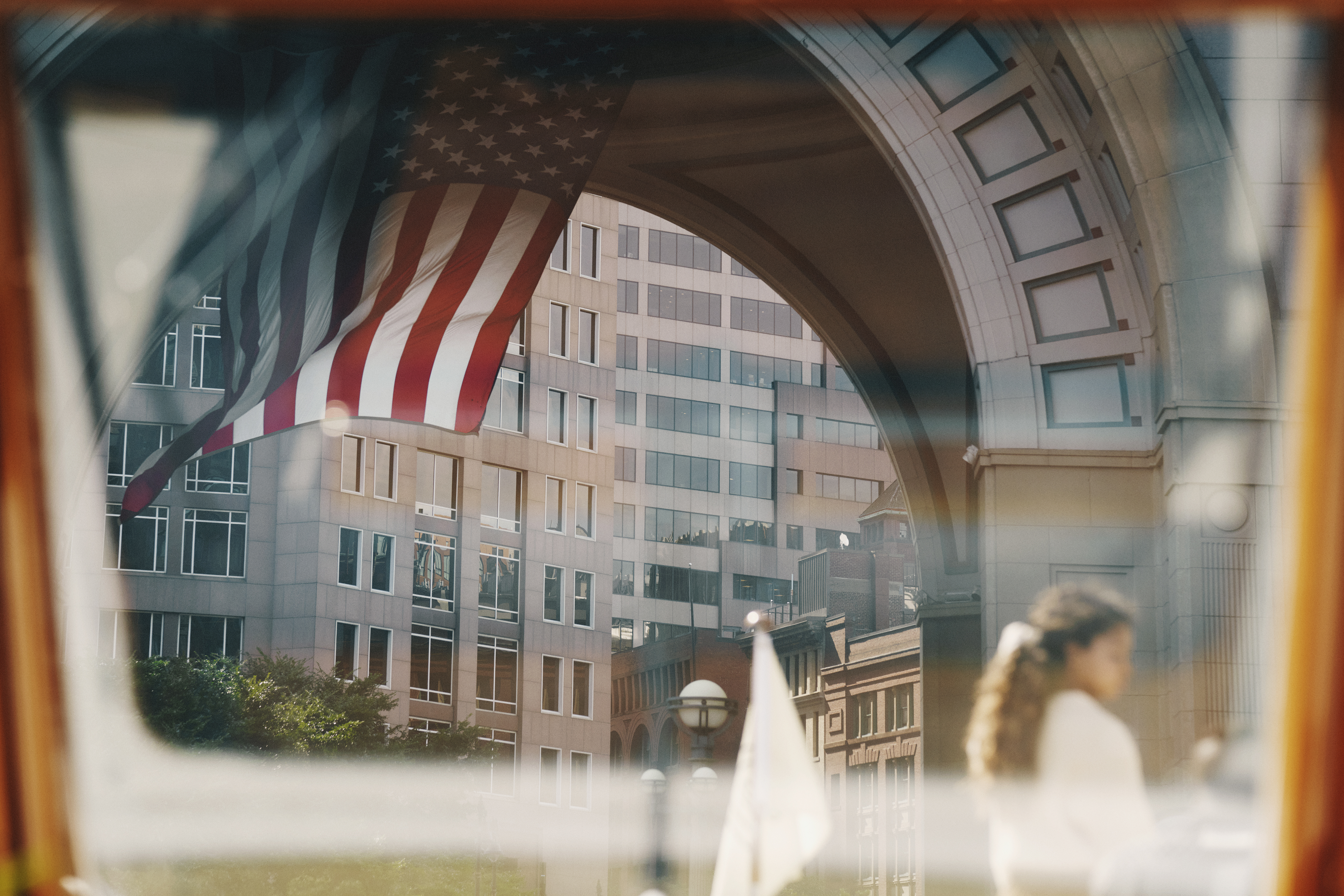 A large American flag hangs inside a grand arched courtyard, framed by white stone buildings and city rooftops beyond.
