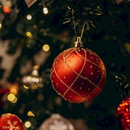 A red jeweled Christmas bauble hangs on a decorated tree, with warm holiday lights softly blurred in the background.