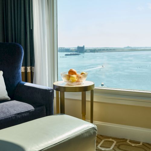 A cozy hotel room with an armchair, ottoman, and a small table with food, overlooking a scenic waterfront view through large windows.