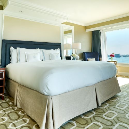 The image shows a well-lit hotel room with a large bed, elegant lamps on side tables, patterned carpet, and a window with a scenic view.