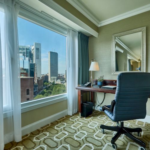 A hotel room with a desk and chair by a large window overlooking a cityscape; a lamp and mirror are on the desk.
