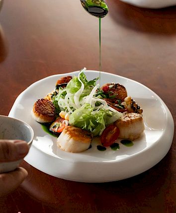 A hand drizzles green sauce over a plated scallop salad as diners eat.