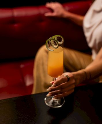 A person sits in a booth holding a golden cocktail with a lime garnish in warm red surroundings.