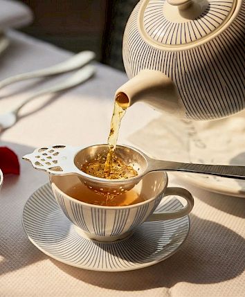 Afternoon tea set on a table with a teapot pouring golden tea into a cup, surrounded by pastries and rose petals.