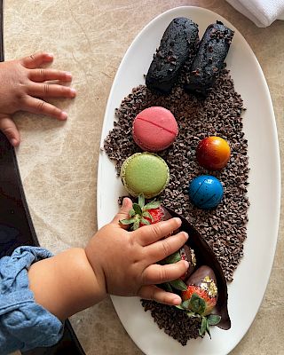 A child's hand reaches for colorful macarons, chocolate truffles, and strawberries on a white plate with chocolate crumbs.