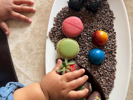 A child's hand reaches for colorful macarons, chocolate truffles, and strawberries on a white plate with chocolate crumbs.