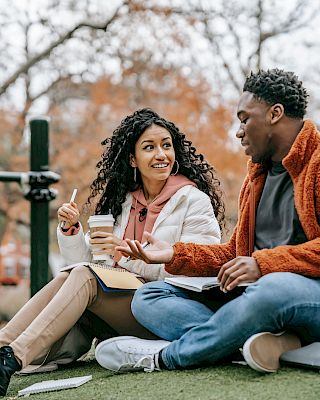 A man and woman sit on grass, talking, with notebooks and a drink, in an outdoor park setting with autumn trees in the background.
