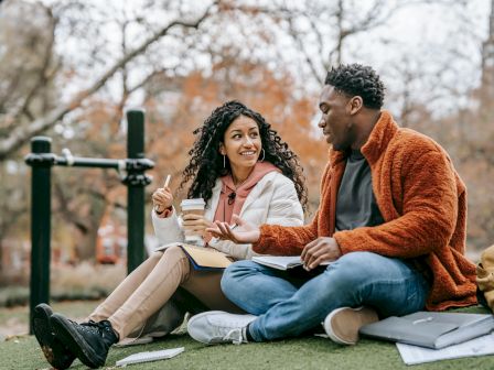 A man and woman sit on grass, talking, with notebooks and a drink, in an outdoor park setting with autumn trees in the background.