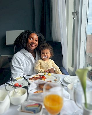 A person and a child are sitting at a table with breakfast items, smiling by a window with light coming through.