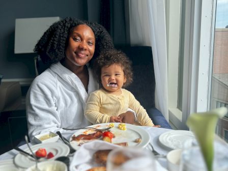 A person and a child are sitting at a table with breakfast items, smiling by a window with light coming through.