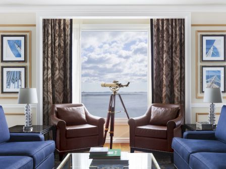 A cozy living room features two armchairs, a telescope by a large window with an ocean view, and blue sofas with lamps and artwork.