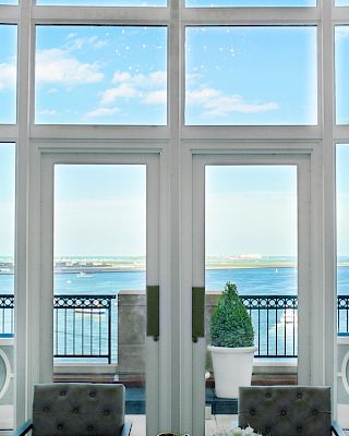The image shows a bright room with large windows overlooking a balcony and a scenic water view, featuring some chairs and a potted plant.