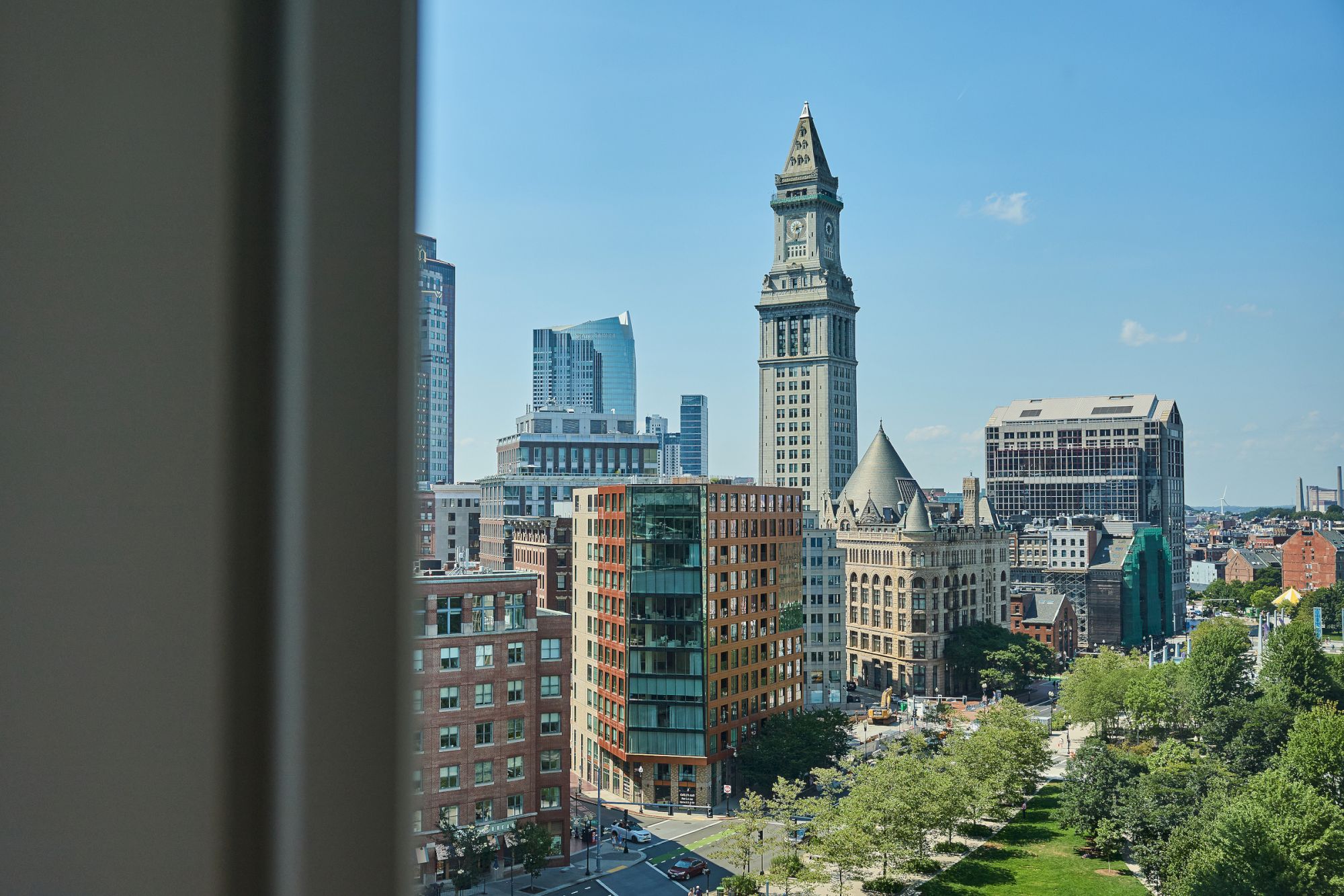 A cityscape featuring a tall clock tower, various modern buildings, and a park with green trees, viewed from a window.