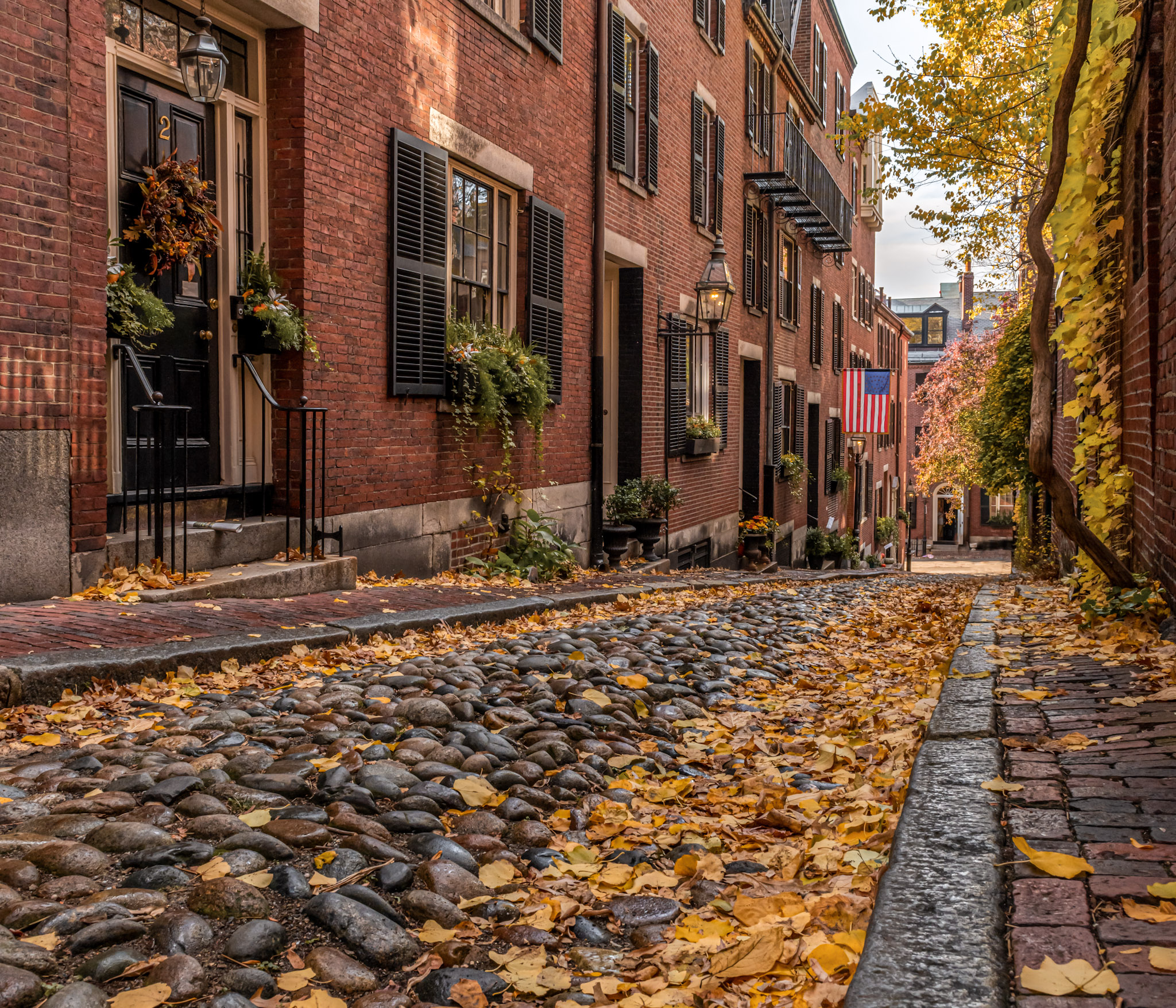 A cobblestone street lined with brick buildings, autumn leaves, and decorated doorways in a quaint neighborhood setting.