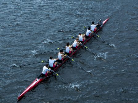 A rowing team in a red boat is moving through the water, with each member synchronized in their strokes.