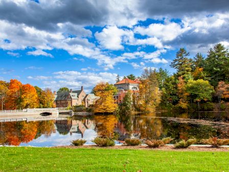 A picturesque autumn scene with colorful trees, a calm lake reflecting the sky and buildings, and a grassy foreground.