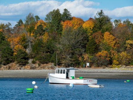 A boat floats on a calm blue lake, surrounded by buoys, with a backdrop of colorful autumn trees and a partly cloudy sky.