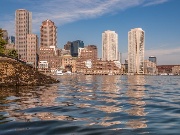 A city skyline lines a river, with tall office towers, a rocky bank, and moored boats under a blue sky.