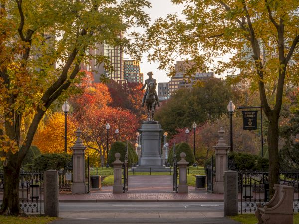A park entrance with a statue centered on the path, surrounded by autumn trees, leading towards the city skyline in the background.
