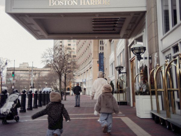 People walk outside a hotel entrance with a marquee reading &ldquo;Boston Harbor,&rdquo; tall buildings in the background, children running toward the street.