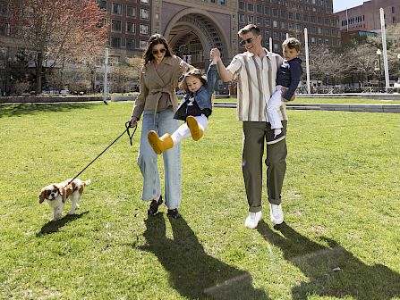 A family strolls on a sunny campus lawn with a small dog and a child, while tall brick buildings stand behind them.