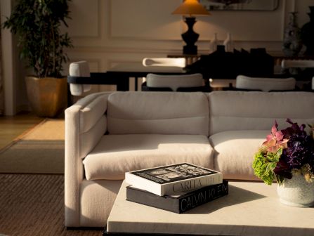 A cozy living room with a cream sofa, coffee table stacked with books, a vase of flowers, and warm lamps.