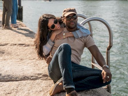 A couple hugs on a sunny waterfront with a city skyline behind them, while another person takes photos nearby.
