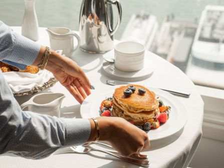 A couple enjoys a sunny brunch: pancakes with berries, a tea pot, cups, and a cream jug on a table by the window.