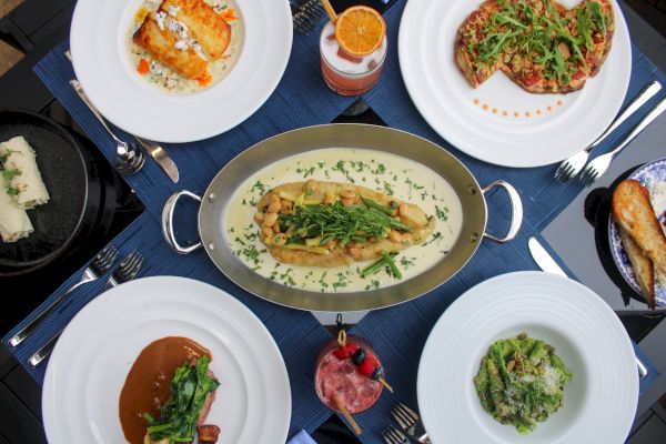 A variety of dishes on a table include pasta, a meat dish, salad, and drinks, all presented on white plates around a central platter.