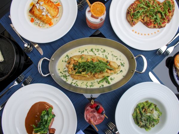 A variety of dishes on a table include pasta, a meat dish, salad, and drinks, all presented on white plates around a central platter.