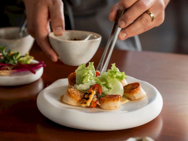 A chef serves seared scallops on a plate, topping them with lettuce and garnish while dining hands wait nearby.