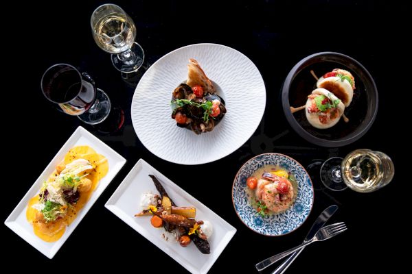 A variety of gourmet dishes with drinks are elegantly arranged on a black surface, featuring different plates and glasses of wine.