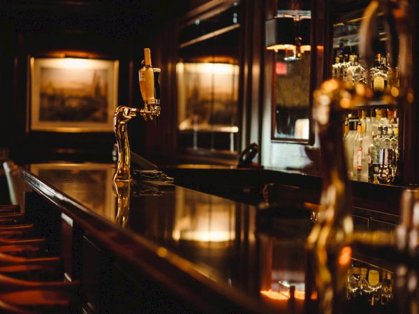 This image shows an empty bar counter with a golden beer tap, wooden bar stools, and shelves stocked with various bottles of alcohol.