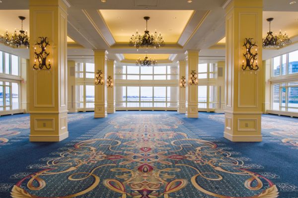Spacious, elegant hall with chandeliers, patterned carpet, large windows showing a water view, and ornate columns.