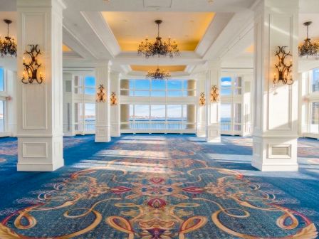 An elegant ballroom with chandeliers, a blue patterned carpet, white columns, and large windows overlooking the sea.
