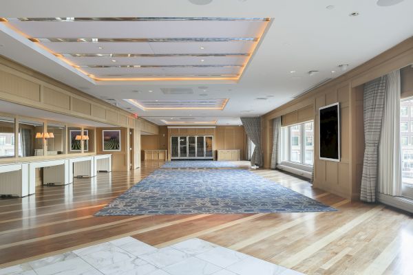 A spacious, elegantly decorated room with a patterned carpet, large windows, wooden floors, and ceiling lights.