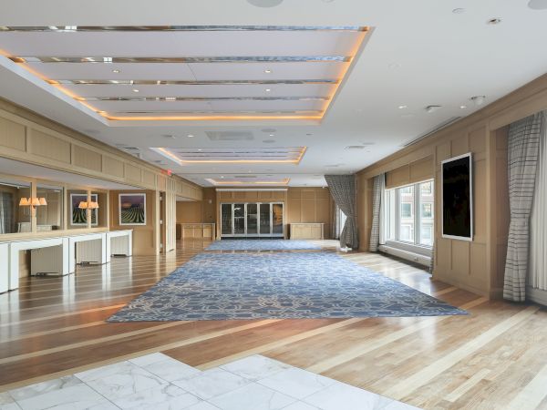 A spacious, elegantly decorated room with a patterned carpet, large windows, wooden floors, and ceiling lights.