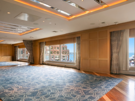 A spacious, well-lit event room with large windows, wooden paneling, patterned carpet, and curtained views of the harbor, ready for meetings.