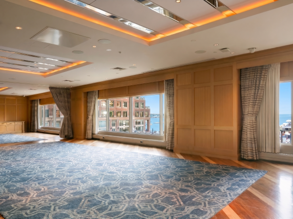 A spacious, well-lit event room with large windows, wooden paneling, patterned carpet, and curtained views of the harbor, ready for meetings.