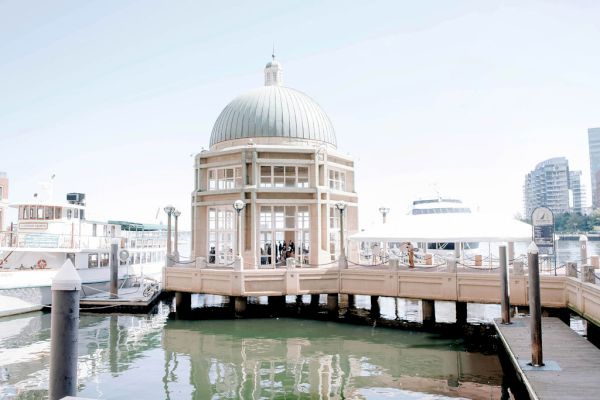 A domed structure by the water, with surrounding boats and nearby tall buildings, under a clear sky.
