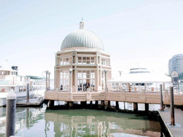 A domed structure by the water, with surrounding boats and nearby tall buildings, under a clear sky.