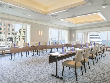 A conference room with U-shaped table setup, chairs, water bottles, and pads, overlooking a waterfront view through large windows.