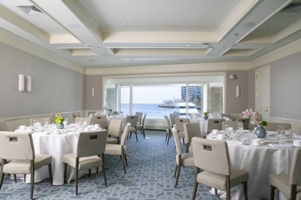 An elegantly set dining room with round tables, flower centerpieces, and a view of water and boats through large windows.